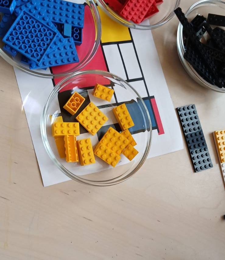 Schalen mit verschiedenen Lego-Steinen, einschließlich gelb, blau und schwarz, stehen auf einem Holztisch. Hinter den Schalen befindet sich ein Mondrian-inspiriertes Gemälde.