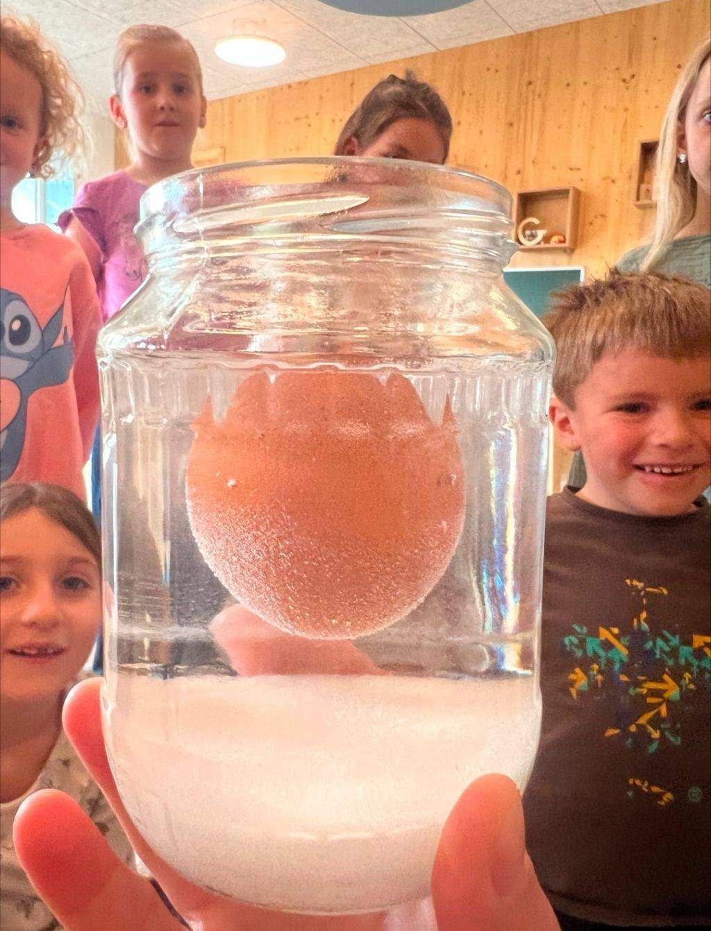 Eine Person hält ein Glas mit einem schwebenden Ei darin, während Kinder dahinter stehen. Das Ei befindet sich in Wasser und scheint zu schweben. Die Kinder tragen Freizeitkleidung.