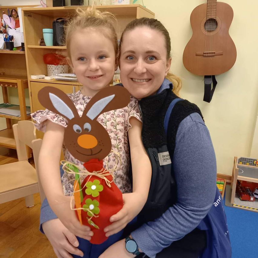 Eine Frau hält ein kleines Mädchen, das einen Hasen-Bastelarbeiten hält, beide lächeln. Hinter ihnen steht ein Bücherregal aus Holz, an der Wand hängt eine Gitarre.