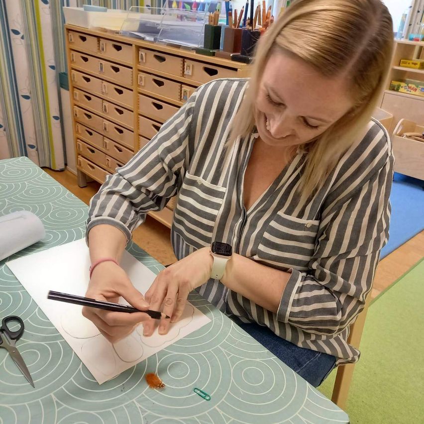 Eine Frau mit blonden Haaren zeichnet auf einem Blatt Papier an einem Tisch, neben ihr liegen Scheren und andere Gegenstände. Hinter ihr steht ein Holzschrank mit vielen Schubladen.