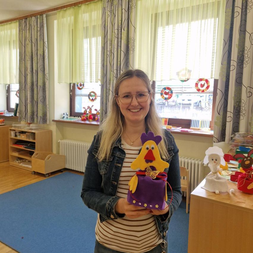 Eine Frau mit Brille und einer Halskette hält ein handgefertigtes Hühnerspielzeug in einem Klassenzimmer mit Holzböden, blauen Vorhängen und einem blauen Teppich.
