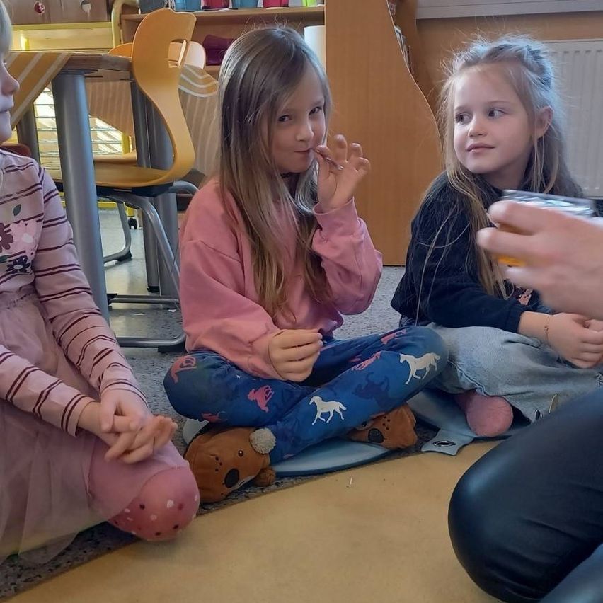 Drei junge Mädchen sitzen auf dem Boden in einem Klassenzimmer, möglicherweise während einer Erzählstunde. Ein Mädchen hält ein Stück Essen in der Hand. Ein anderes Mädchen hat einen Becher in der Hand. Ein Plüschtier liegt neben dem Mädchen in der Mitte.