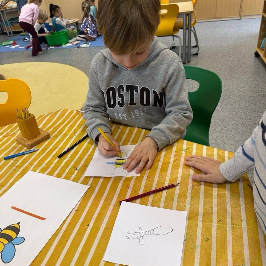 Ein junger Junge zeichnet eine Biene auf Papier mit einem Bleistift. Er sitzt an einem Tisch mit anderen Kindern.