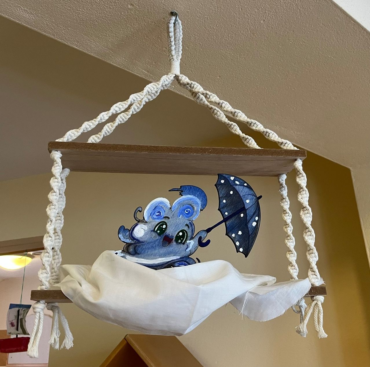 Ein verspielter blauer Elefant mit einem Regenschirm hängt von der Decke mit einem weißen Stoffbett. Der Elefant ist auf einer hölzernen Plattform gemalt, die von weißen Seilen aufgehängt ist.