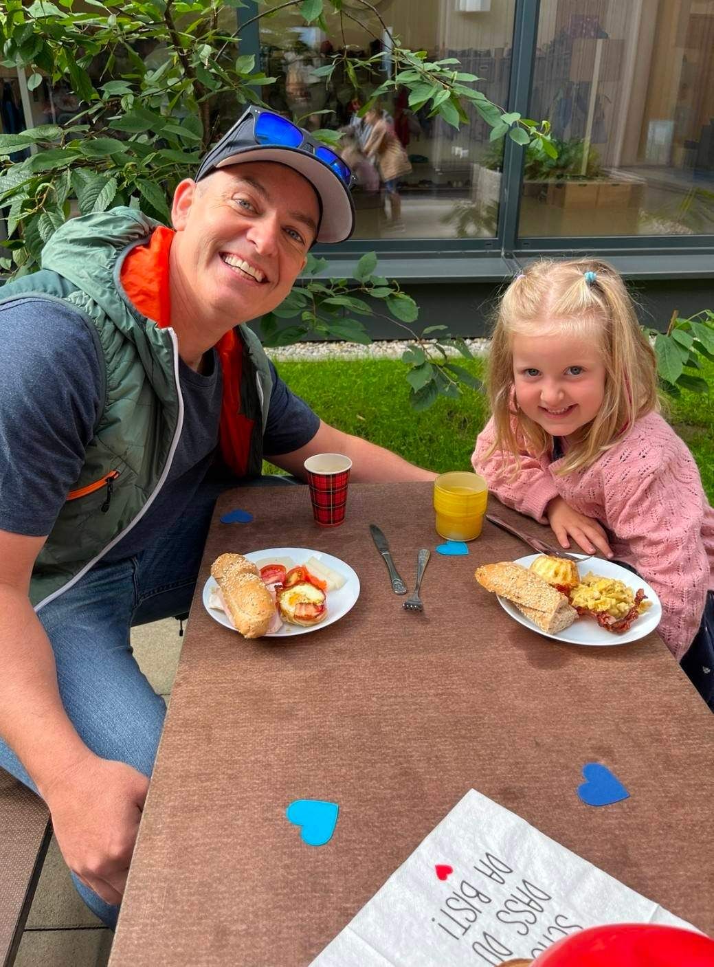 A man and a girl sit at a table, smiling, with breakfast in front of them, including a sandwich, eggs, and juice.