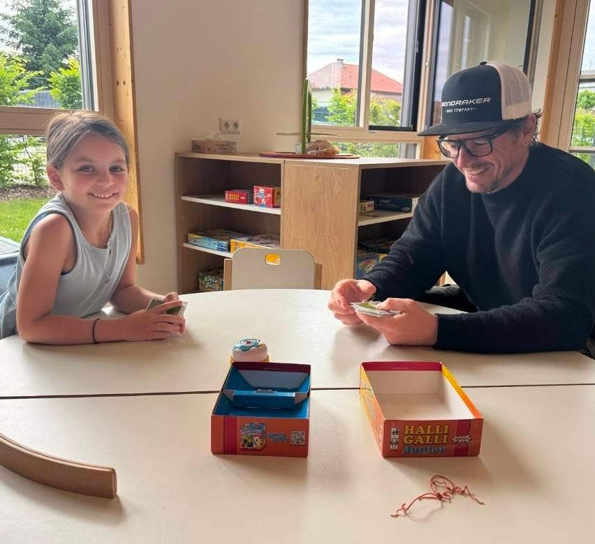 An adult man and a young girl are playing a board game at a table, smiling. The game box and cards are on the table, and a shelf with various items is behind them.