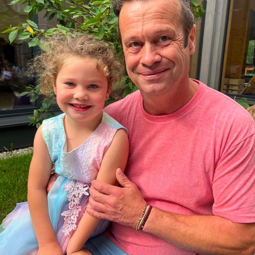 A man in a pink shirt holds a smiling young girl in a pink dress, sitting on his lap in a garden.