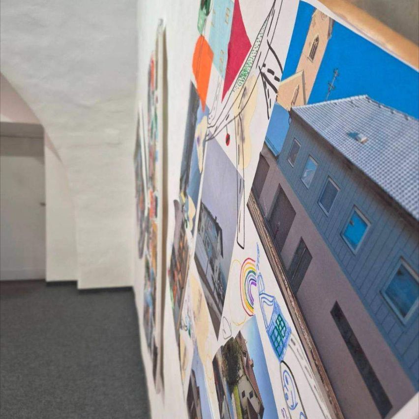 Ein Korridor mit einer weißen Wand, die mit verschiedenen farbigen Collagen verschiedener Gebäude, eines Regenbogens und einer Kirche verziert ist. Der Boden ist mit Teppich ausgelegt und die Decke ist weiß.