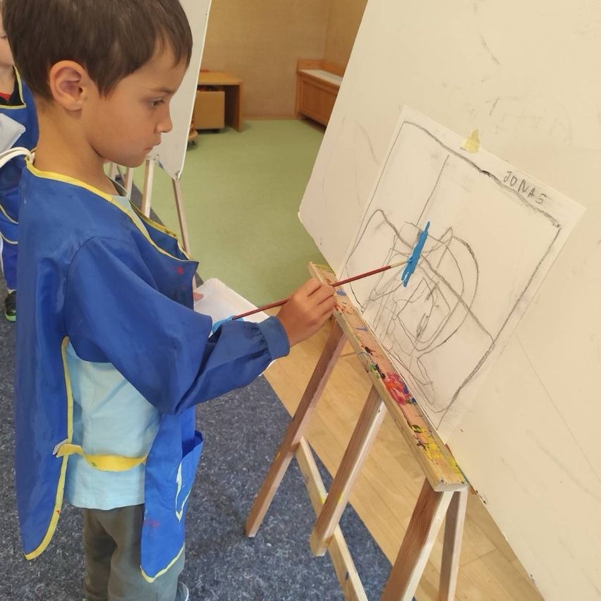 Ein Junge in einem blauen Malerkittel malt auf einer Leinwand in einem Klassenzimmer. Es gibt andere Schüler im Hintergrund.