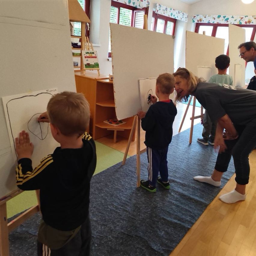 Zwei Jungen zeichnen mit Hilfe eines Erwachsenen auf große weiße Tafeln. Andere Kinder sind im Hintergrund.
