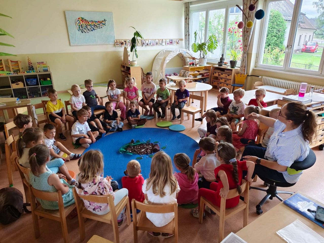 Viele kleine Kinder sind in einem Kreis um einen Erwachsenen versammelt, wahrscheinlich in einem Klassenzimmer. Ein blauer Teppich mit kuchenartigem Design liegt auf dem Boden.