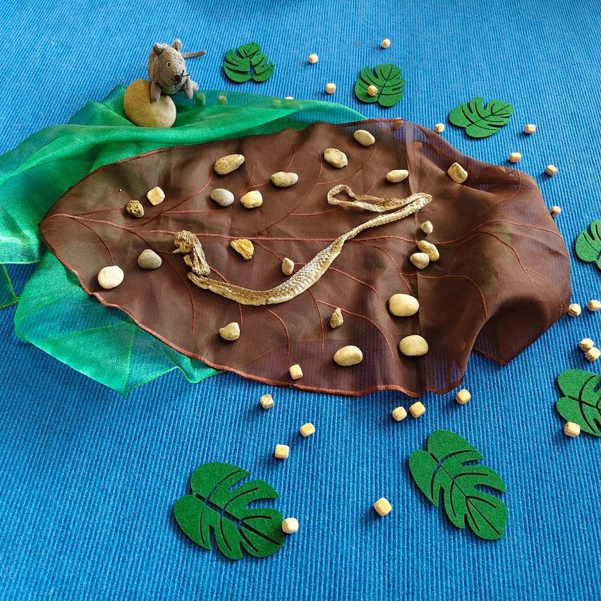 Ein Blatt mit einer Schlange und Steinen auf einem blauen Teppich mit einem kleinen Plüschtier und verstreuten grünen Blättern.