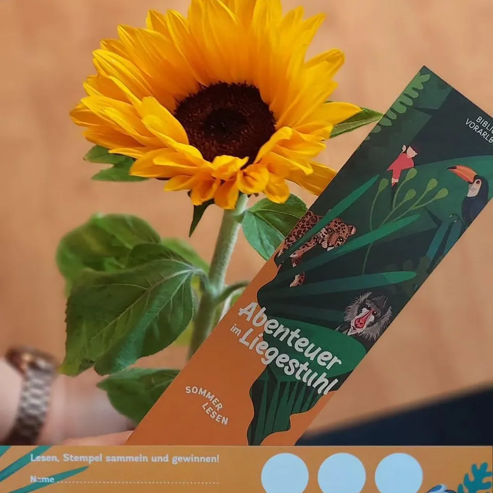 Ein Lesezeichen mit dem Titel 'Abenteuer im Liegestuhl' mit einer Dschungelszene wird neben einer Sonnenblume gehalten. Das Lesezeichen ist braun und grün, mit einem weißen Kreis am unteren Rand.