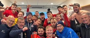 Eine Gruppe von Fußballspielern in roten und blauen Trikots feiert in der Umkleidekabine, mit Bierflaschen und einem Feuerlöscher in der Hand.