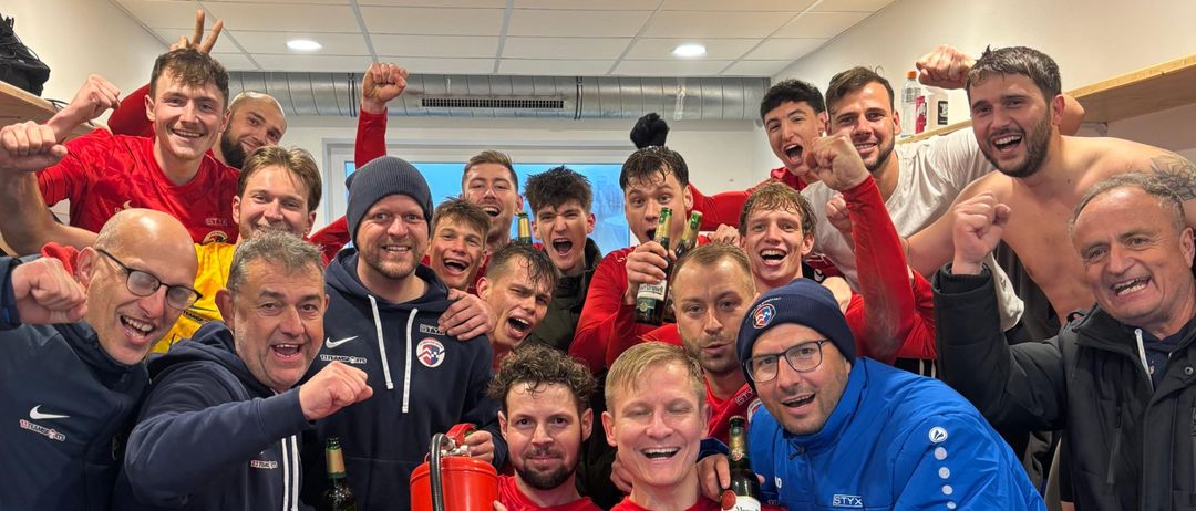 Eine Gruppe von Fußballspielern in roten und blauen Trikots feiert in der Umkleidekabine, mit Bierflaschen und einem Feuerlöscher in der Hand.
