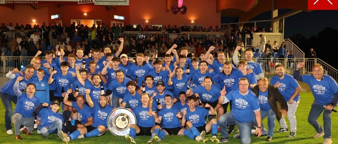Eine Fußballmannschaft feiert mit einer Trophäe auf einem Feld, umgeben von Fans in einem Stadion. Die Spieler tragen blaue Trikots mit 'Meister' darauf.