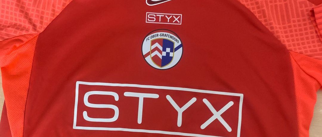 Ein rotes Fußballtrikot mit Nike-Logo und einem weißen STYX-Logo auf der Vorderseite. Das Trikot zeigt auch ein Teamlogo.