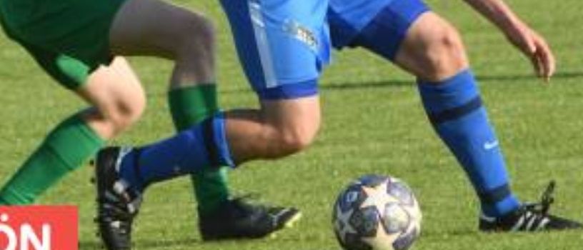 Zwei Fußballspieler in blauen und grünen Trikots spielen ein Spiel auf einem Feld. Der Spieler in Blau tritt den Ball, während der andere versucht, ihn zu blockieren.