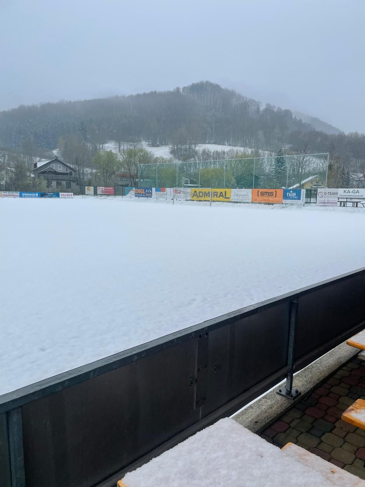 Snow-covered sports field with a fence, distant hills, and trees. Banners on the fence include Admiral, SMS, and Thir.