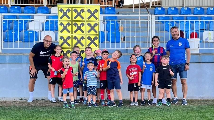 A group of children and two adults stand on a soccer field, smiling and holding ice pops. The children wear various jerseys, and a yellow board with black Xs is behind them. Empty blue bleachers are in the background.