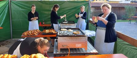 Frauen bereiten und braten Lebensmittel an einem Stand unter einem Vordach. Eine Frau beugt sich vor, um das Essen auf einem Tablett zu untersuchen.