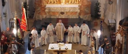 Eine religiöse Zeremonie findet in einer Kirche mit mehreren Personen statt. Altäre mit Kerzen, Blumen und anderen Dekorationen schmücken die Kirche. Eine Gruppe von Menschen steht vor dem Altar und hält sich an den Händen.