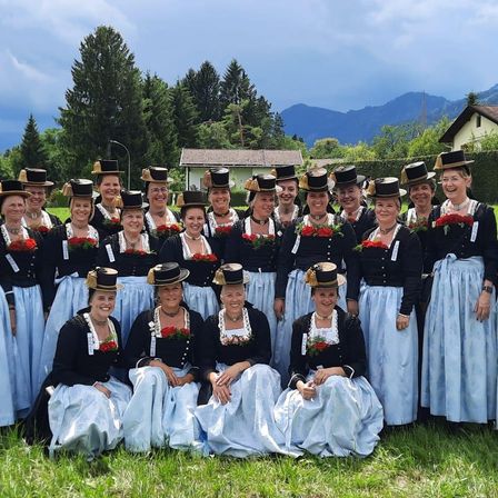 Eine Gruppe von Frauen in traditioneller Kleidung, einschließlich schwarzer Oberteile und blauer Röcke, posiert für ein Foto auf einem Rasenfeld mit Bergen im Hintergrund.