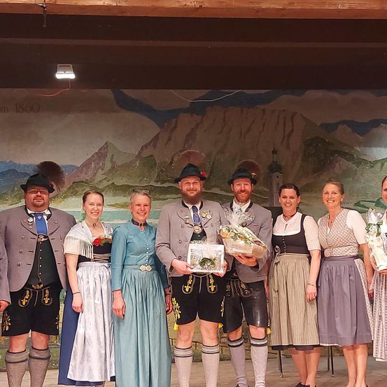 Gruppe von Menschen in traditioneller bayerischer Kleidung, die auf einer Bühne stehen und Blumensträuße und Auszeichnungen halten. Dahinter zeigt eine Wandmalerei eine Berglandschaft und ein Gebäude.