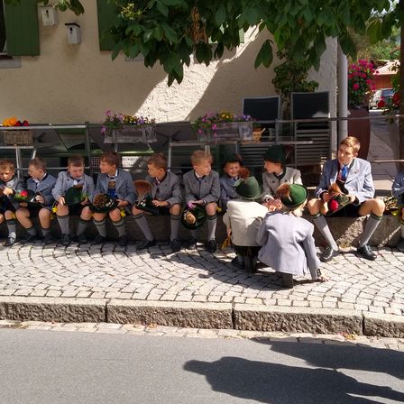 Eine Gruppe von Kindern in grünen Hüten sitzt auf einem Bordstein. Sie tragen traditionelle Kleidung und halten Blumen. Ein Gebäude mit grünen Fensterläden und hängenden Pflanzen steht hinter ihnen.
