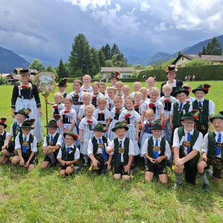 Eine Gruppe von Kindern und Erwachsenen in traditioneller Kleidung posiert für ein Foto auf einer Wiese mit Bergen im Hintergrund.