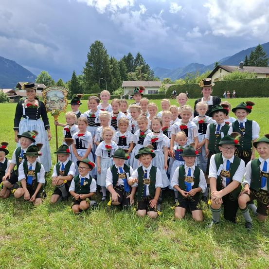 Eine Gruppe von Kindern und Erwachsenen in traditioneller Kleidung posiert für ein Foto auf einer Wiese mit Bergen im Hintergrund.