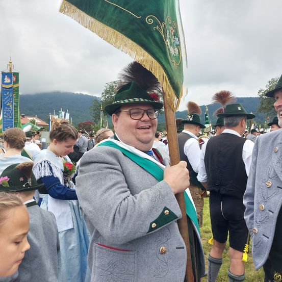 Ein Mann in traditioneller bayerischer Kleidung hält eine grüne Flagge mit einer Feder bei einem Fest. Ihn umgeben Menschen in traditioneller Kleidung, und Berge sind im Hintergrund.