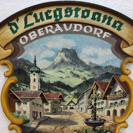 Ein gemaltes Schild, das das Dorf Oberaudorf mit einer Kirche und Bergen im Hintergrund zeigt.