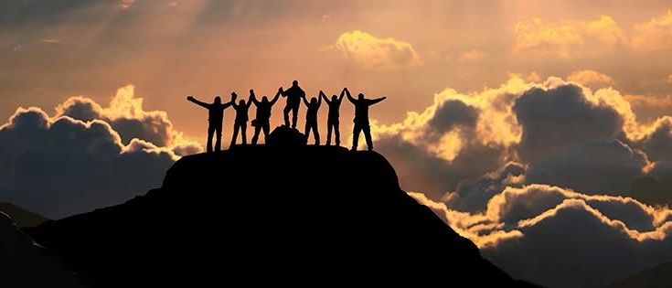 Eine Gruppe von Menschen in Silhouette mit erhobenen Armen in Triumphstellung, die auf einem Berggipfel steht, vor einem Himmel voller Wolken und einer untergehenden Sonne.