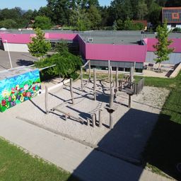 Eine Draufsicht auf einen Spielplatz mit Holzgeräten in einem Sandbereich, mit einem bunten Wandbild auf der linken Seite und Gebäuden mit rosa Dächern im Hintergrund.