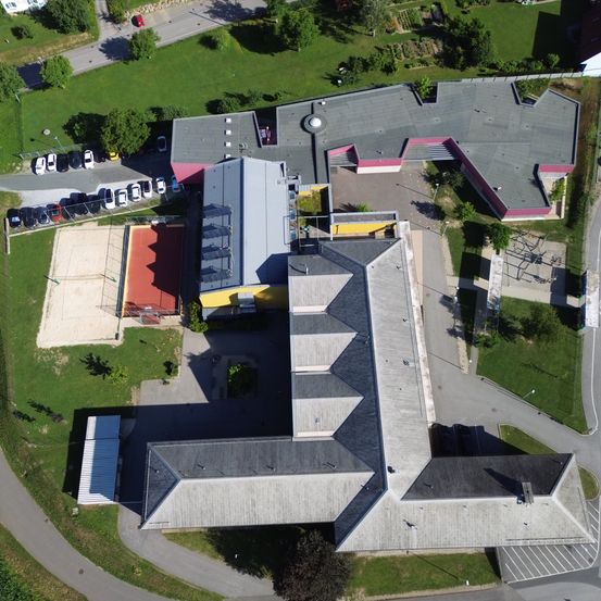 Ein Luftbild einer Schule mit mehreren Gebäuden, einem Spielplatz und einem Parkplatz. Die Gebäude haben graue Dächer und rosa Akzente.