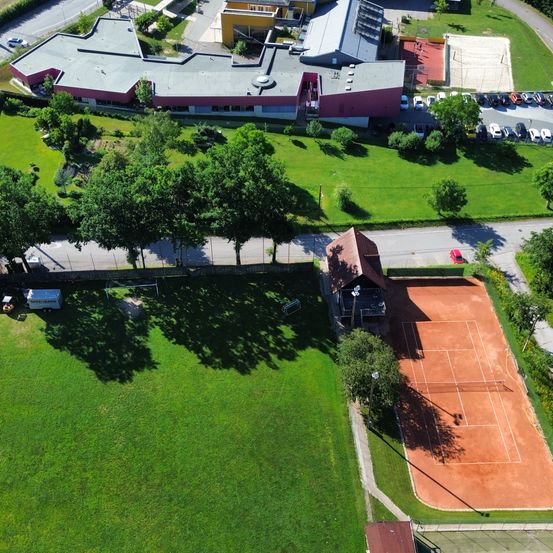 Luftaufnahme eines Sportkomplexes mit einem Tennisplatz, einem Spielplatz und einem großen Gebäude mit rotem Dach. Mehrere Autos sind auf dem Parkplatz abgestellt.