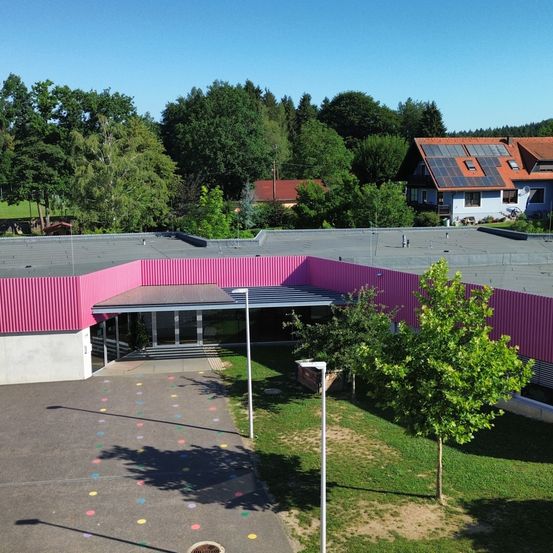 Ein Luftbild eines Gebäudes mit rosa Außenwand und grauem Dach, umgeben von Bäumen. Das Gebäude befindet sich in einem Grasbereich mit bunten Punkten. Ein Haus mit Solarmodulen auf dem Dach ist in der Ferne.
