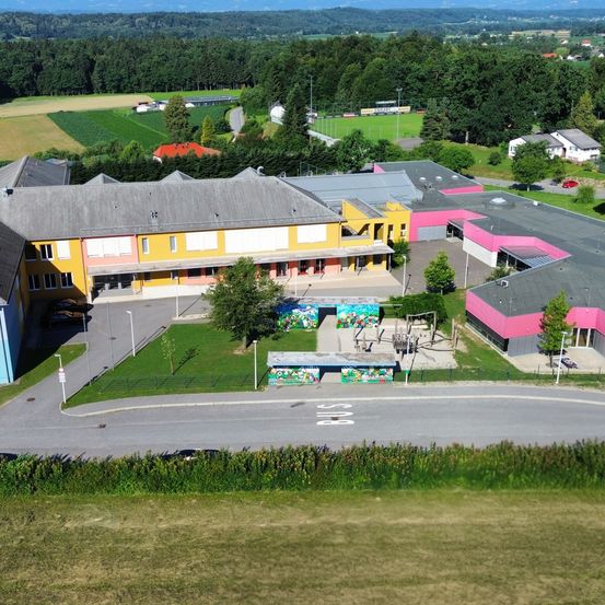 Luftaufnahme einer Schule mit gelben und rosa Gebäuden. Sie ist umgeben von einem grünen Feld, Bäumen und Häusern. Es gibt einen Spielplatz und ein kleines Häuschen mit bunten Designs.