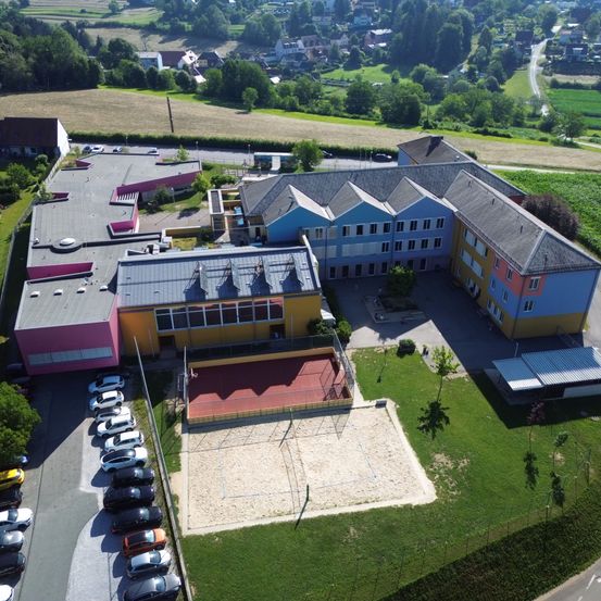 Ein Luftbild einer Schule mit mehreren Gebäuden, einem roten Tennisplatz, einem Sandvolleyballplatz und vielen Autos, die davor geparkt sind.