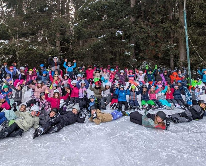 Eine große Gruppe von Menschen posiert für ein Foto auf einer Eisbahn mit verschneiten Bäumen im Hintergrund.