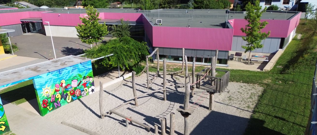 Ein Spielplatz mit einem hölzernen Hindernisparcours steht vor einem Gebäude mit einem rosa Dach. Es gibt Bäume und eine Wandmalerei.