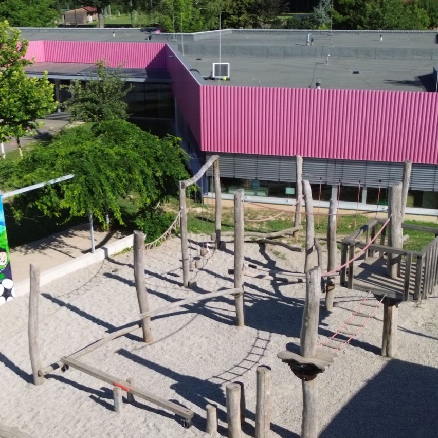 Ein Spielplatz mit einem hölzernen Hindernisparcours steht vor einem Gebäude mit einem rosa Dach. Es gibt Bäume und eine Wandmalerei.