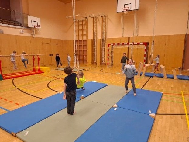 Eine Gruppe von Kindern spielt auf dem Hallenboden mit Matten und Reifen. Ein Basketballkorb und ein Tor sind an der Wand angebracht.