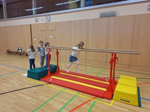 Vier Kinder trainieren in einer Turnhalle. Ein Kind ist auf der Reckstange, während die anderen zuschauen. Die Turnhalle hat Holzböden, braune Wände und eine Uhr an der Wand.
