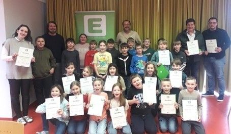 Eine Gruppe lächelnder Kinder und Erwachsener posiert für ein Foto und hält Urkunden in den Händen. Die Kinder sitzen und stehen vor einem Hintergrund mit dem Buchstaben E.