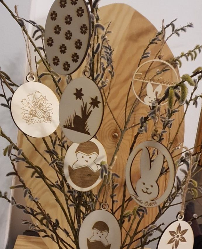 Ein Holzbrett zeigt eine Sammlung von Oster-Themen-Holzschmuck. Die Ornamente umfassen Designs von Blumen, Sternen, Eiern und Hasen. Jedes Ornament ist einzigartig verziert und verleiht der Ausstellung eine festliche Note.