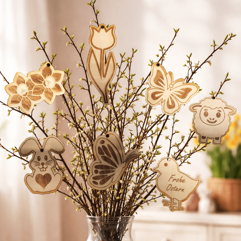 Eine Glassvase mit Frühlingszweigen, die mit hölzernen Osterdekorationen wie einem Hasen, einer Tulpe, einem Schmetterling und einem Schaf verziert sind. Die Vase zeigt auch einen Vogel mit dem Text 'Frohe Ostern'.