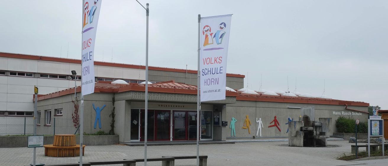 Außenansicht der Volksschule Horn mit Bannern, Bänken und einem Straßenlicht davor. Das Gebäude hat ein rot-oranges Dach und farbenfrohe Figuren an den Wänden.