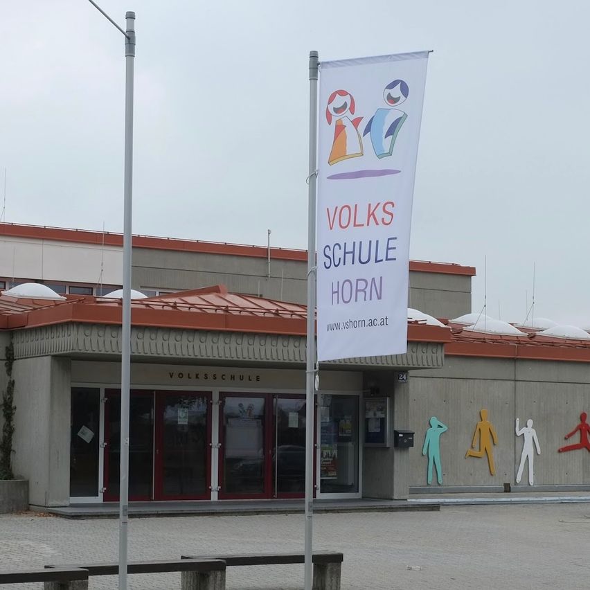Außenansicht der Volksschule Horn mit Bannern, Bänken und einem Straßenlicht davor. Das Gebäude hat ein rot-oranges Dach und farbenfrohe Figuren an den Wänden.
